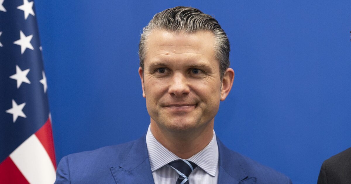 War Secretary Pete Hegseth, in front of a blue backdrop and an American flag.