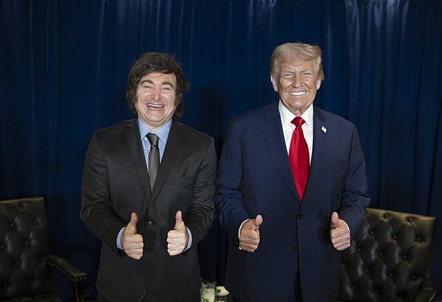 Two men giving thumbs up pose together, one in a suit and tie and the other in a black suit, against a blue curtain backdrop.