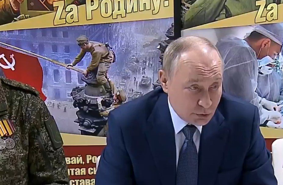 Russian leader Vladimir Putin speaking at a meeting, with military personnel and historical imagery in the background highlighting themes of patriotism and sacrifice.