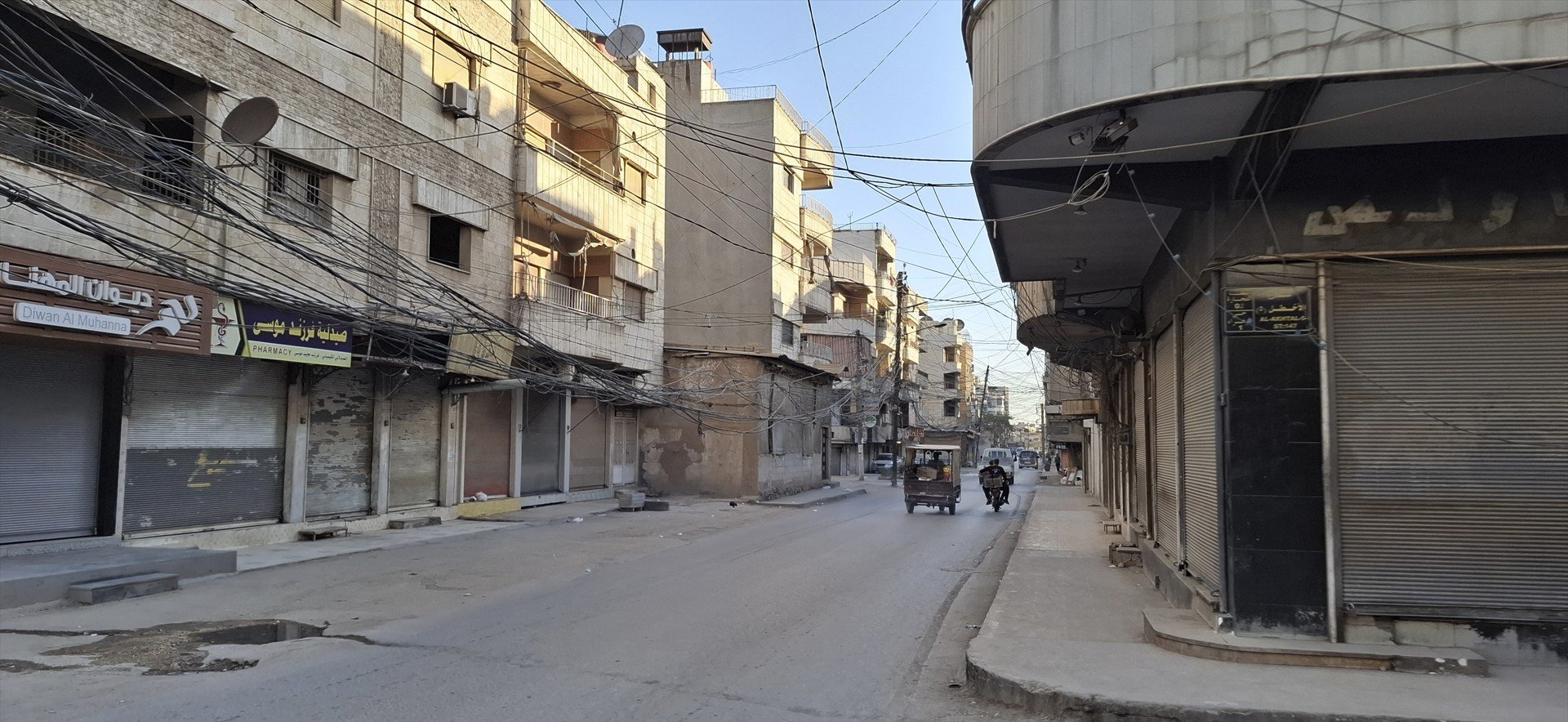 Empty street scene in an urban area with buildings, shops, and tangled overhead wires during daylight.