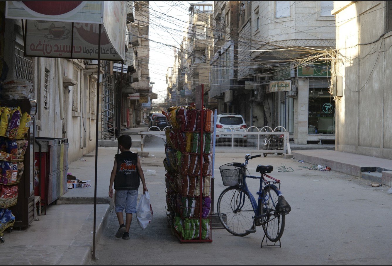 A child walks down a quiet street lined with shops and a parked bicycle, capturing a moment of daily life in an urban setting.