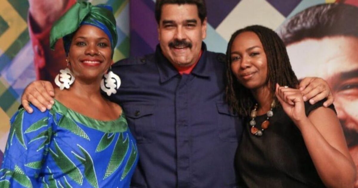 Nicolas Maduro poses with two women, one in traditional attire, at a vibrant event backdrop featuring geometric patterns.