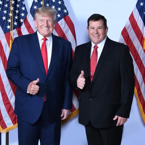 Donald Trump and a supporter pose together with thumbs up in front of American flags, highlighting a moment of camaraderie at a political event.