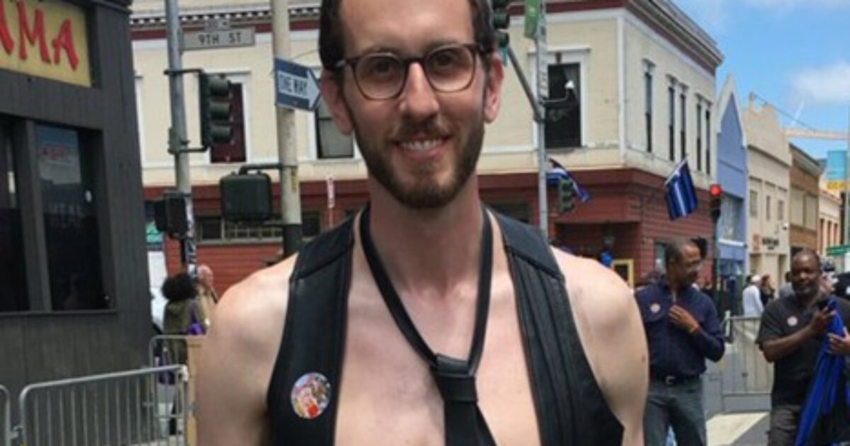 Man wearing a harness and glasses smiles at a street event, with a crowd and buildings in the background.