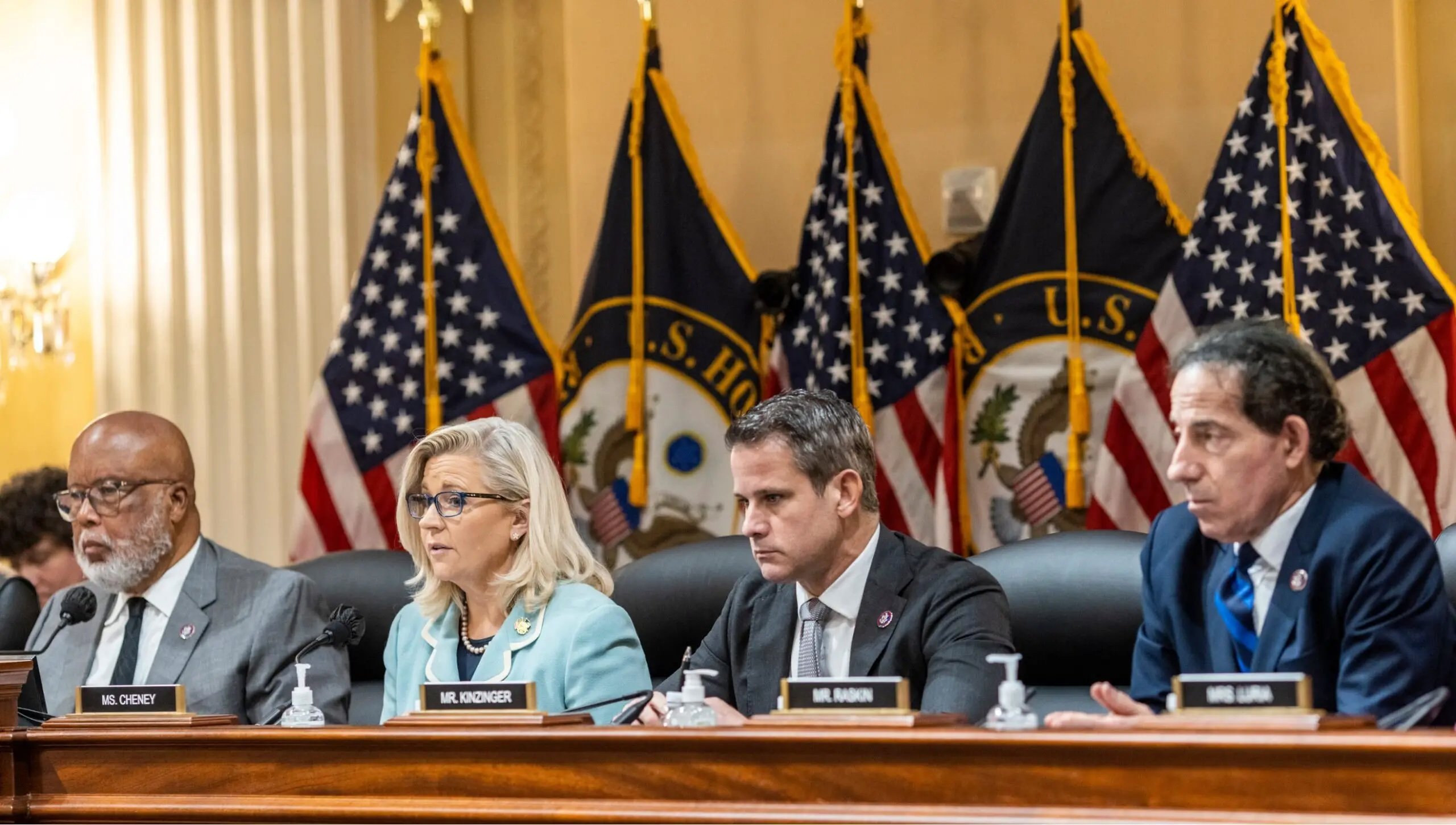 Members of a congressional committee seated at a table, discussing important issues, with multiple U.S. flags in the background.