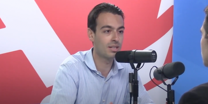Man in a light blue shirt speaking into a microphone during a podcast interview, with a colorful background featuring red and blue elements.