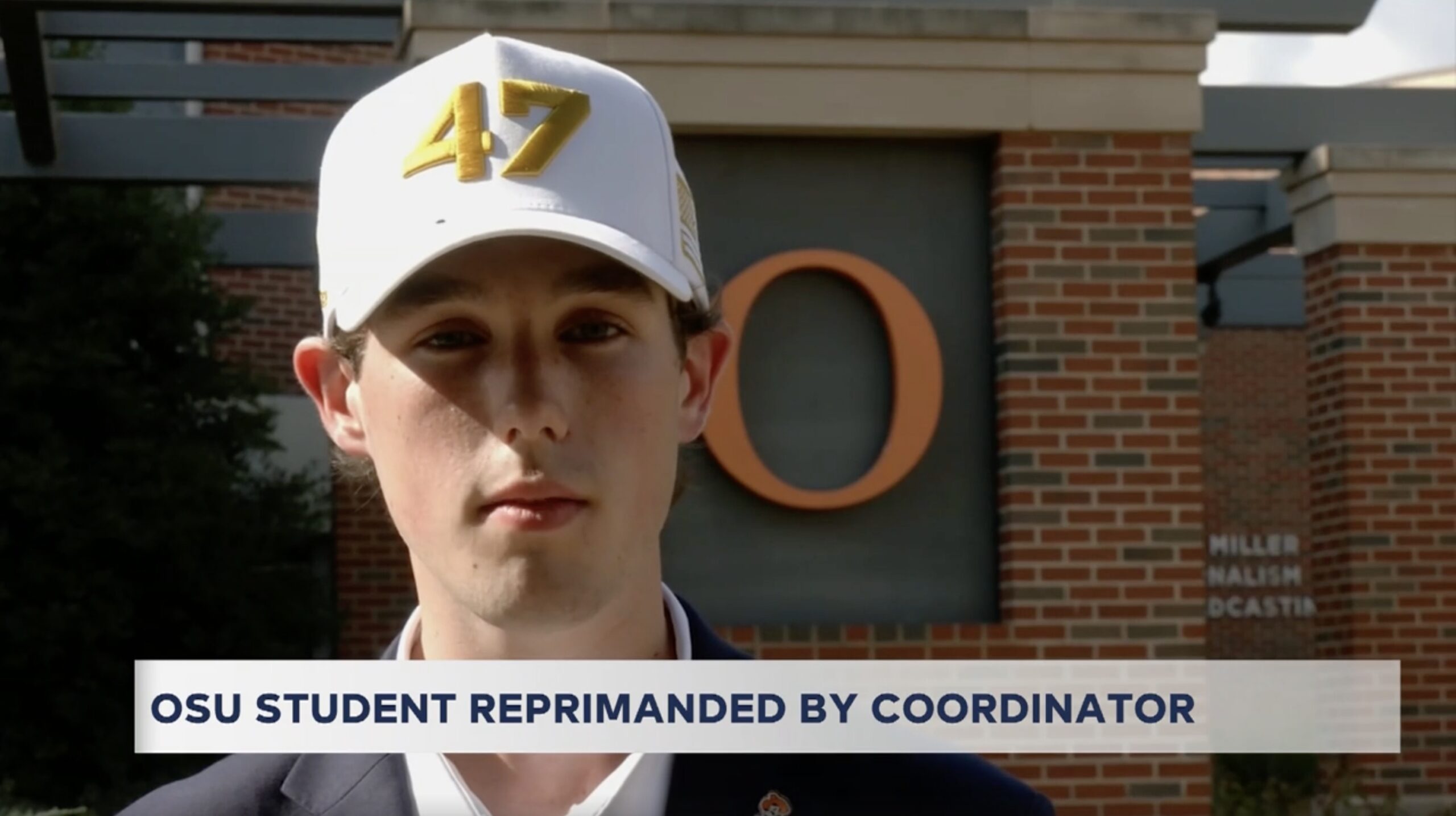 OSU student wearing a white cap with the number 47 stands in front of a university building, discussing a recent reprimand by a coordinator.