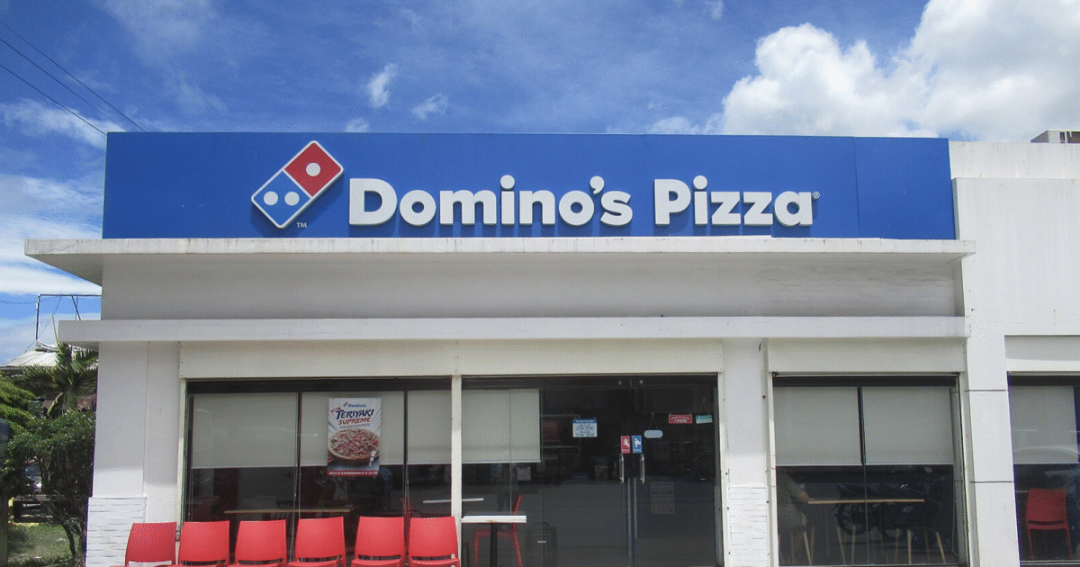 Domino's Pizza storefront featuring a blue sign, red chairs outside, and a clear sky backdrop, ideal for takeout and dining options.