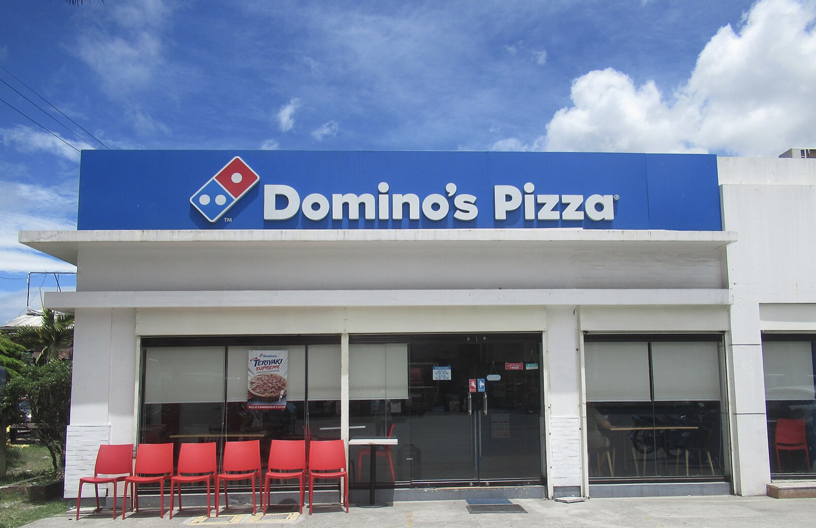Domino's Pizza storefront featuring a blue sign, red chairs outside, and a clear sky backdrop, ideal for takeout and dining options.