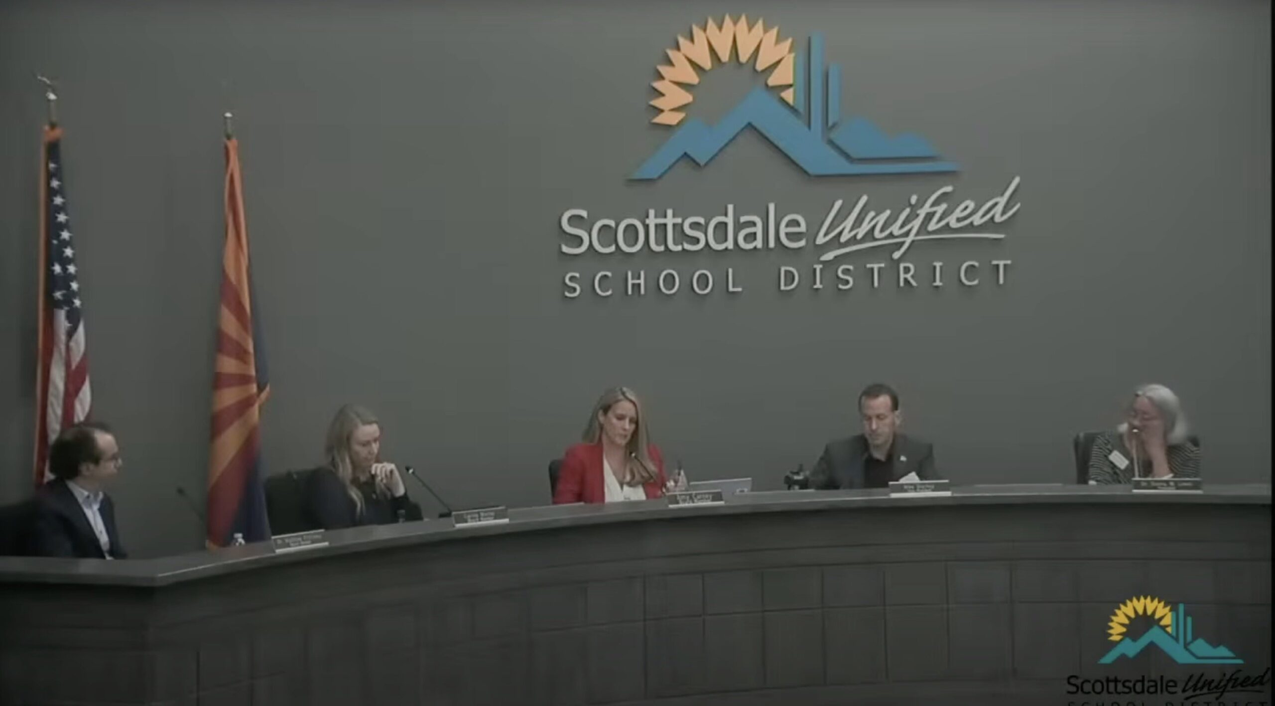 Scottsdale Unified School District board members discuss educational policies during a meeting, with flags displayed in the background.