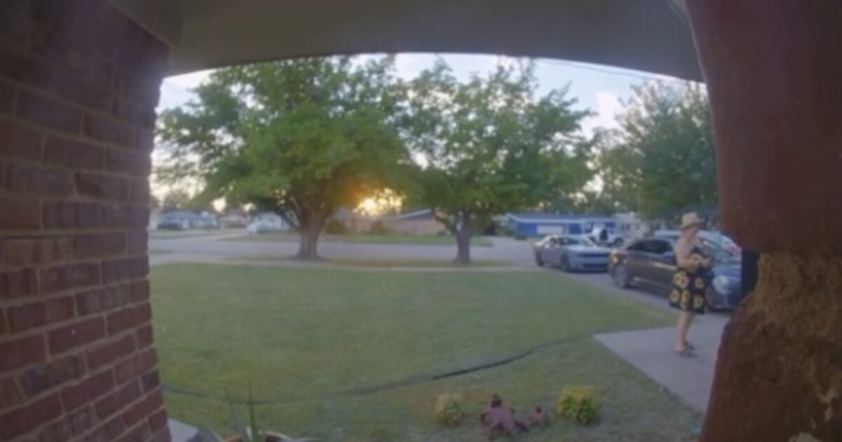 A woman in a floral dress stands on a porch at sunset, with trees and parked cars visible in the background.
