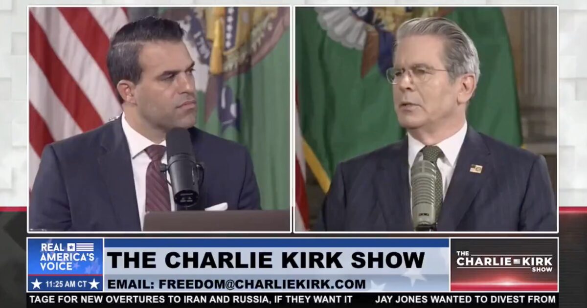 Two men engage in discussion on The Charlie Kirk Show, featuring a backdrop of flags and a microphone setup.
