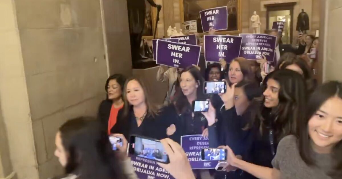 A group of women holding signs advocating for the swearing-in of Adelita Grijalva, demonstrating in a public building.