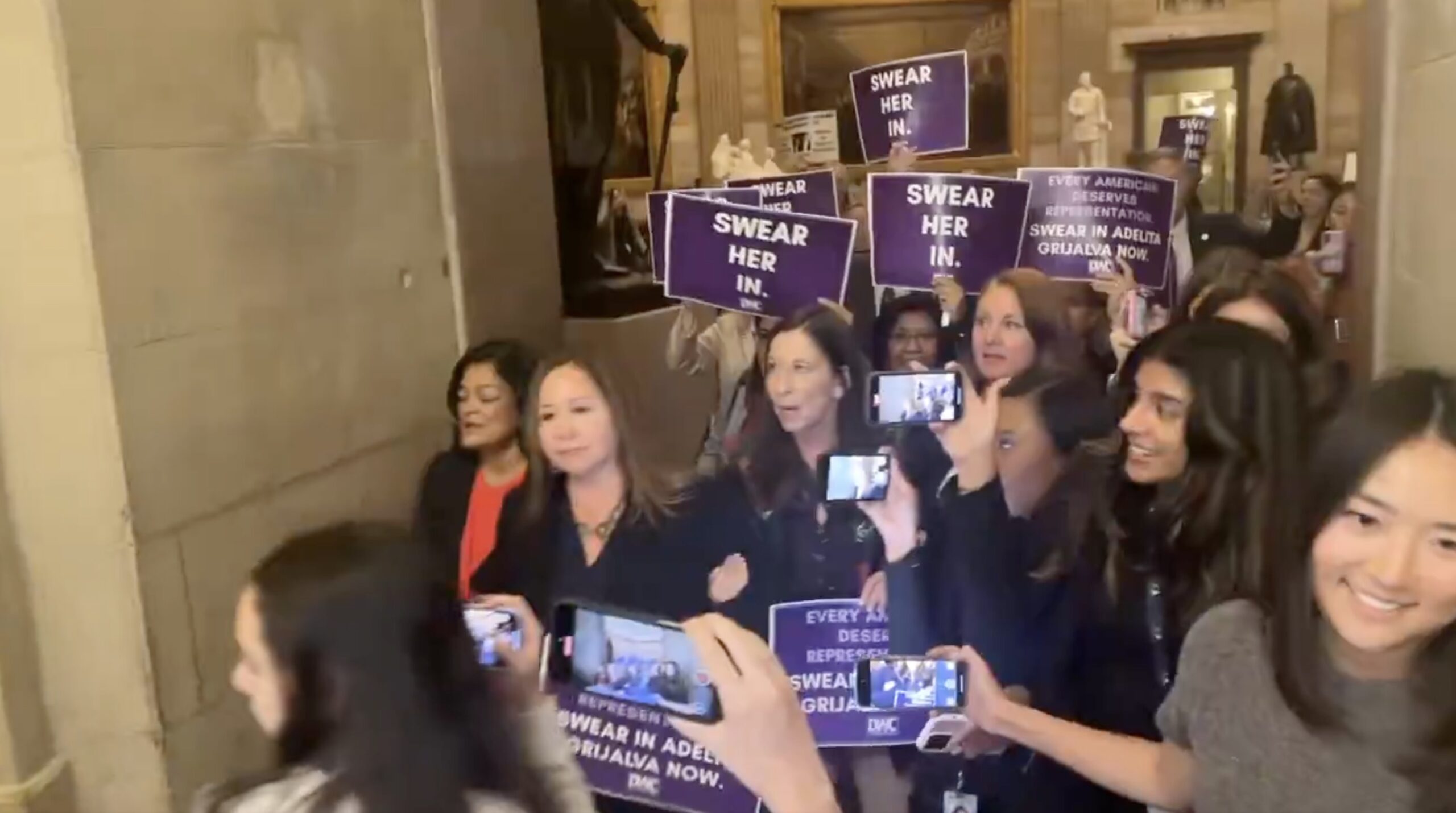 A group of women holding signs advocating for the swearing-in of Adelita Grijalva, demonstrating in a public building.