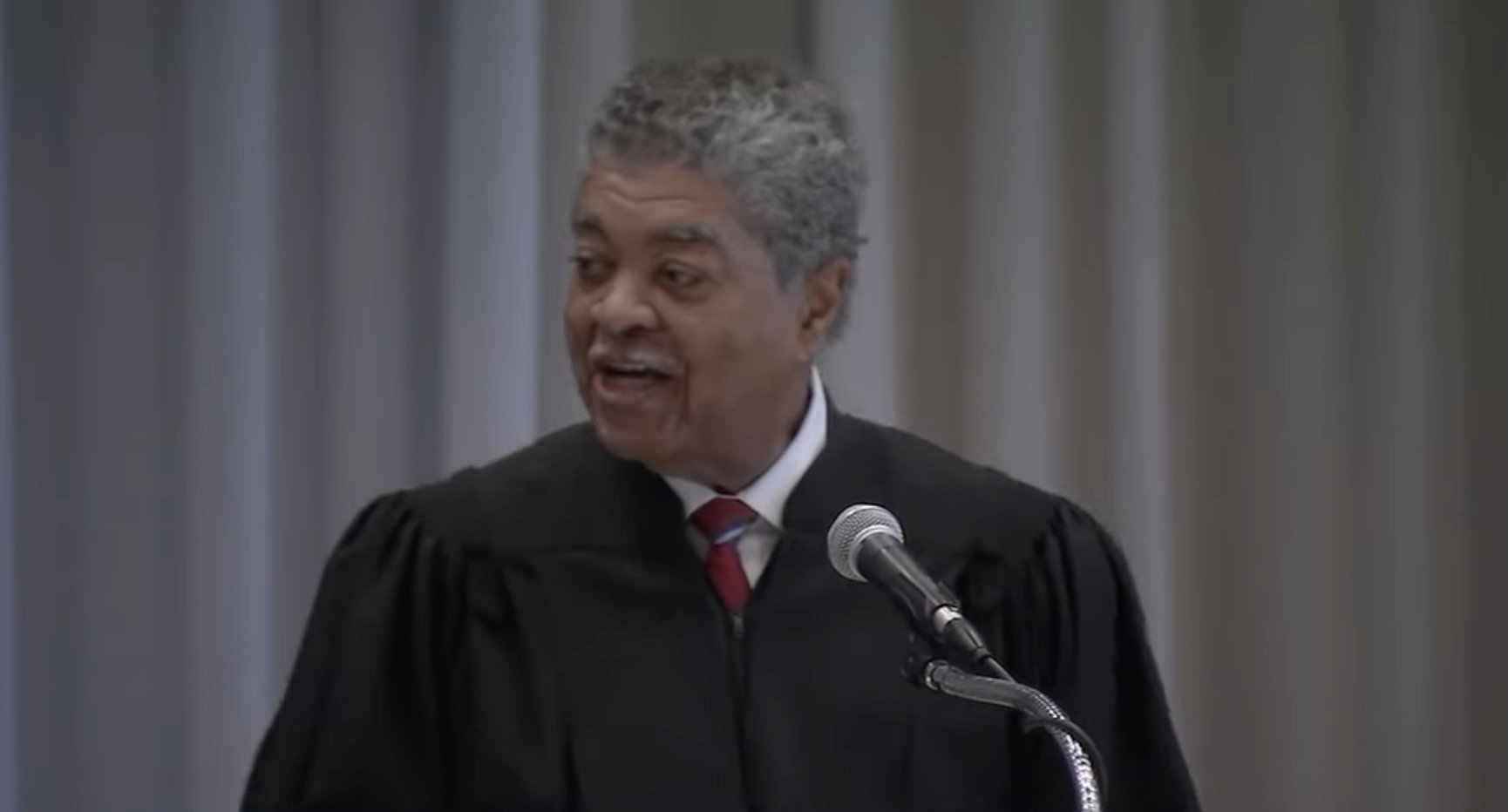 Judge speaking at a formal event, wearing a black robe and delivering an engaging speech at a podium with a microphone.