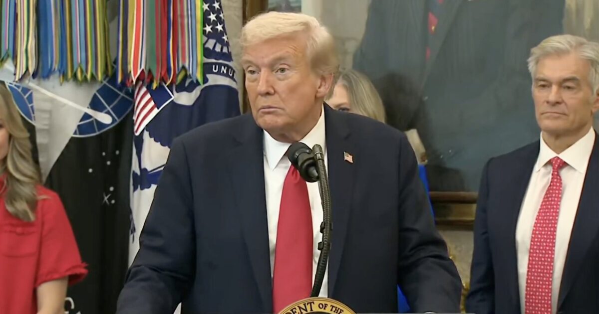 Donald Trump speaks at a podium in front of military decorations, flanked by supporters during a press event.