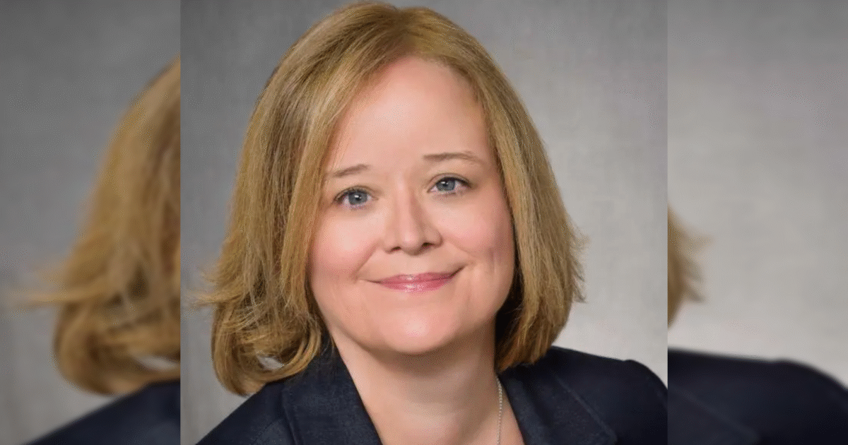 Professional headshot of a woman with shoulder-length blonde hair, smiling, wearing a dark blazer against a neutral background.