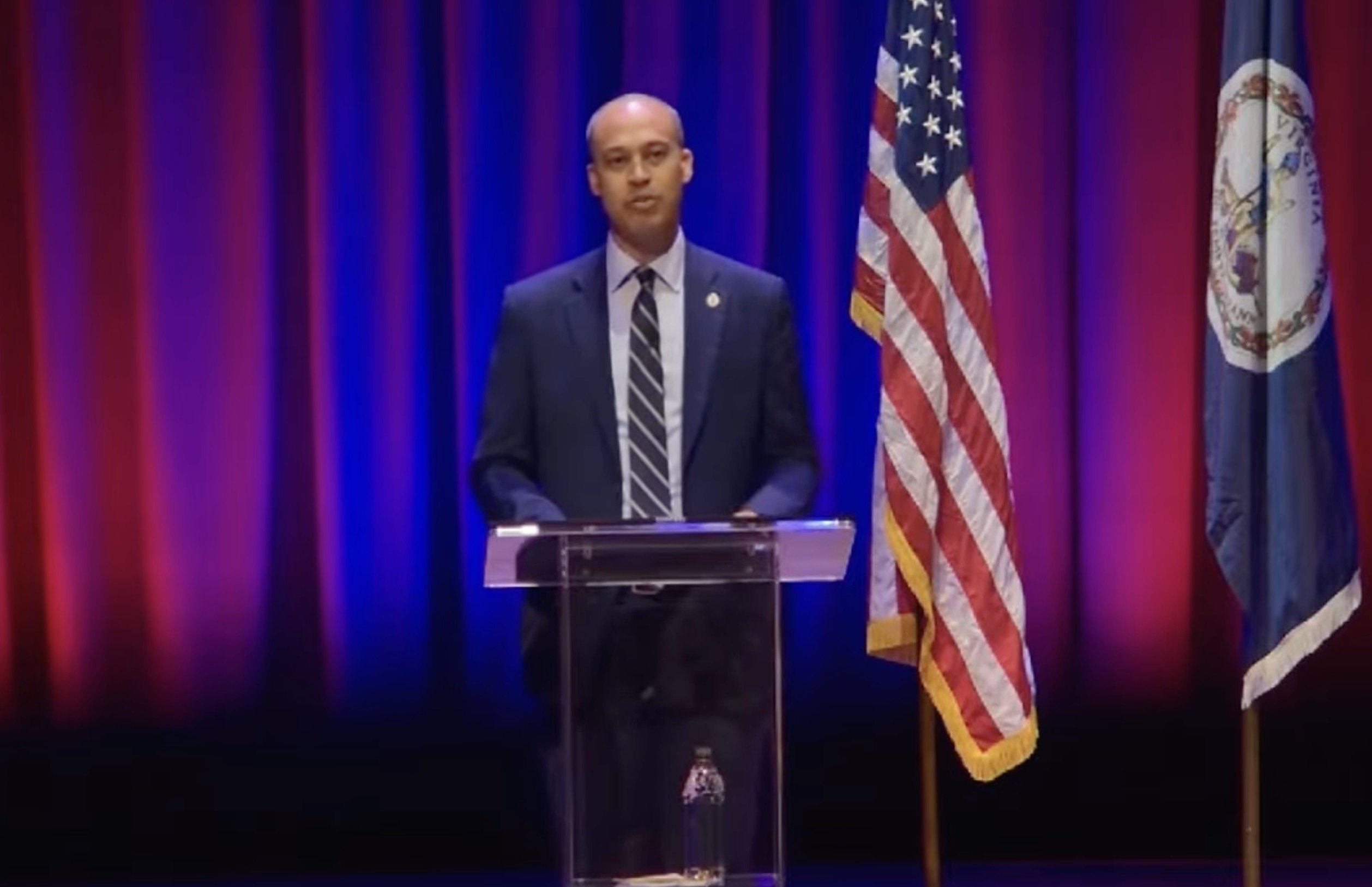 A speaker stands at a podium with American and Virginia flags in a well-lit auditorium, addressing an audience during a formal event.