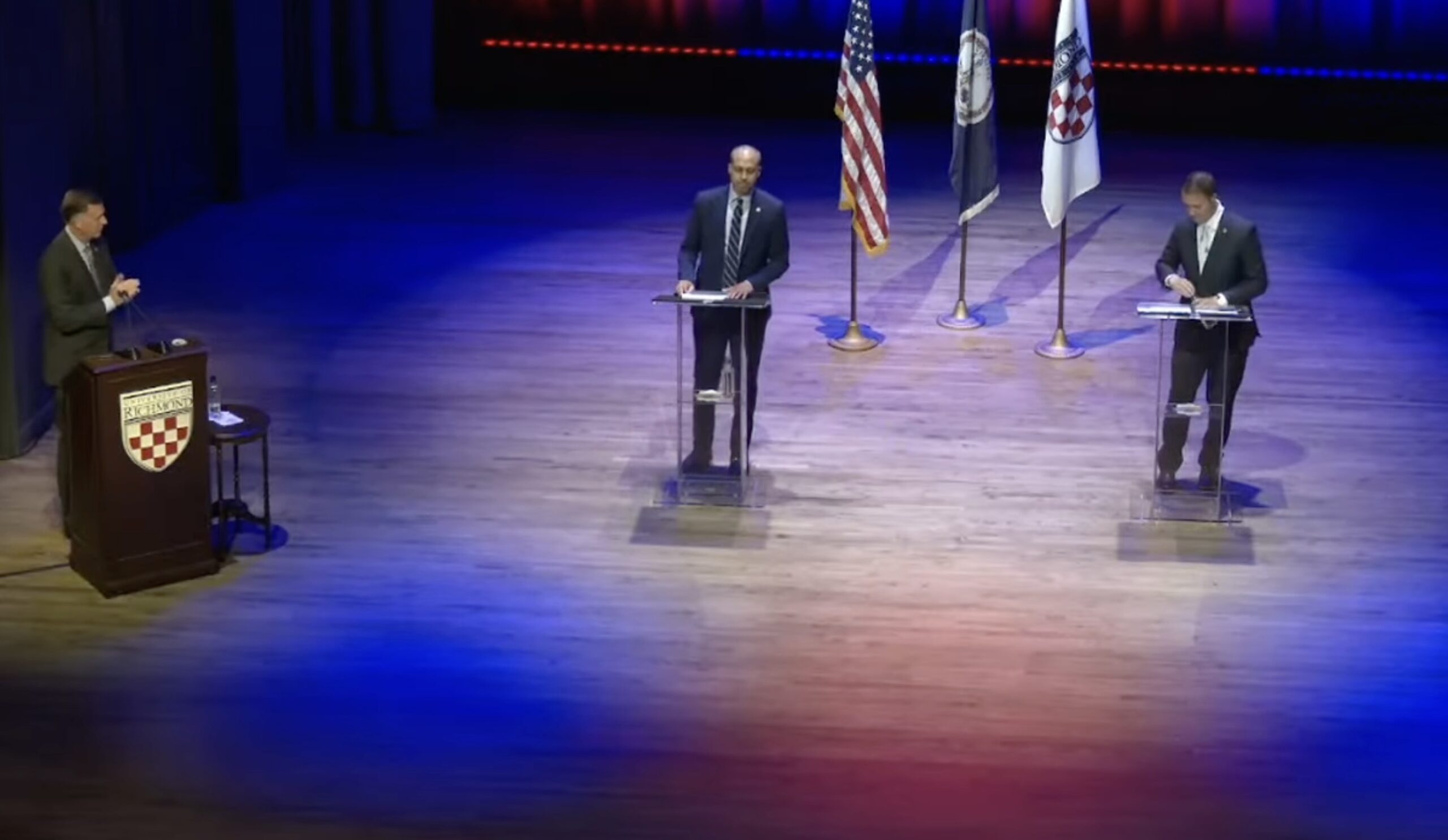 Candidates participating in a political debate on stage with flags in the background and a moderator at a podium.