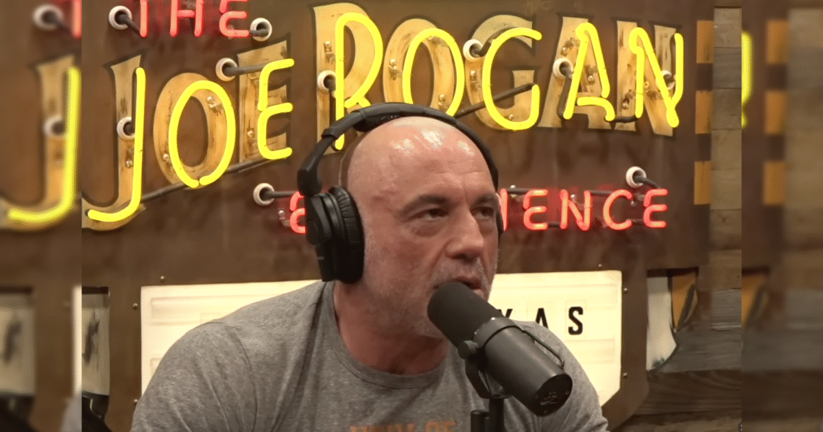 Joe Rogan speaking into a microphone during a podcast episode, with colorful neon signs in the background promoting "The Joe Rogan Experience."