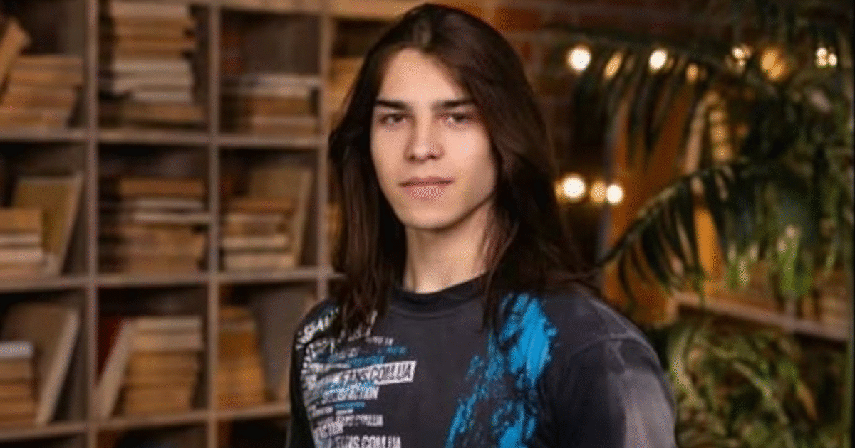 Young man with long hair posing confidently in front of a bookshelf, wearing a black shirt with blue graphics.