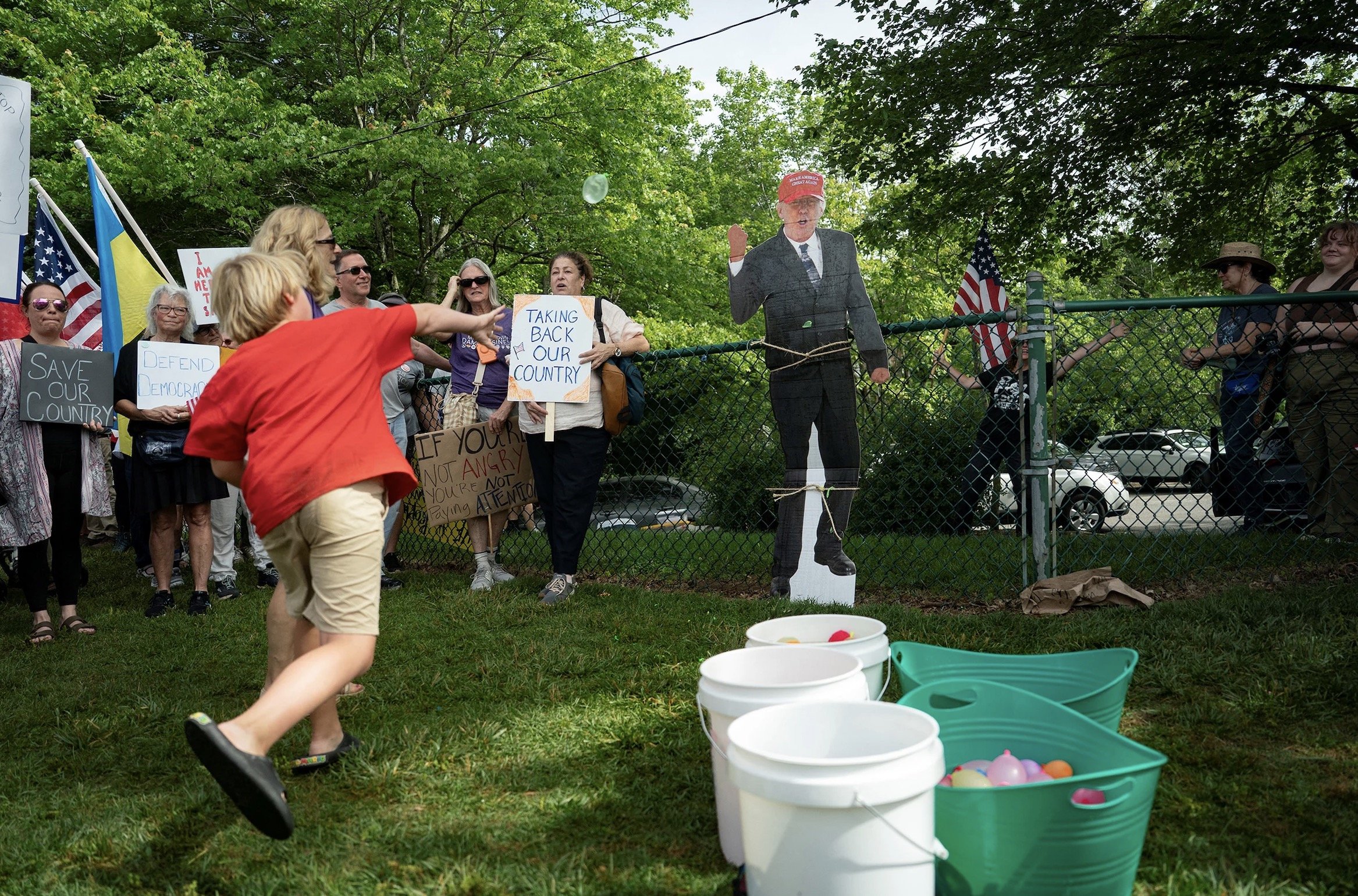 A child throws a water balloon at a cardboard cutout of a politician while protesters hold signs advocating for democracy and political change in a park setting.