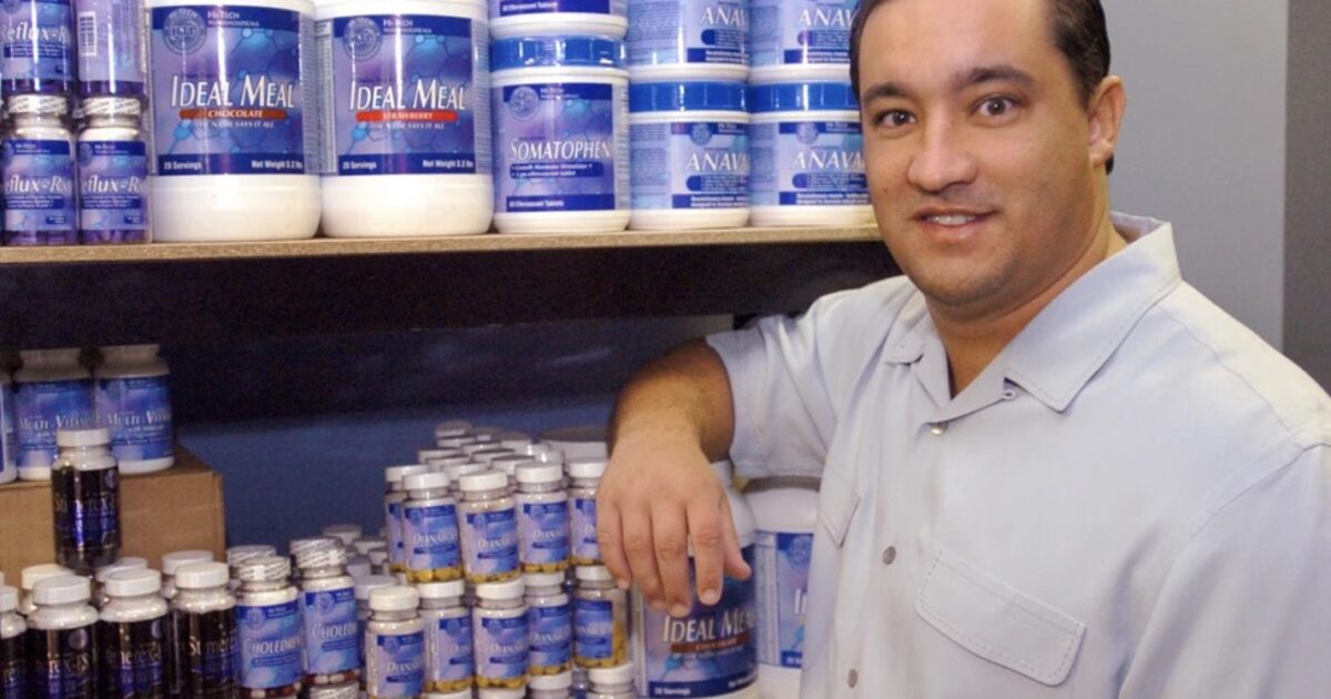 Man standing next to shelves filled with various nutritional supplements and protein powders, showcasing a health and wellness store environment.