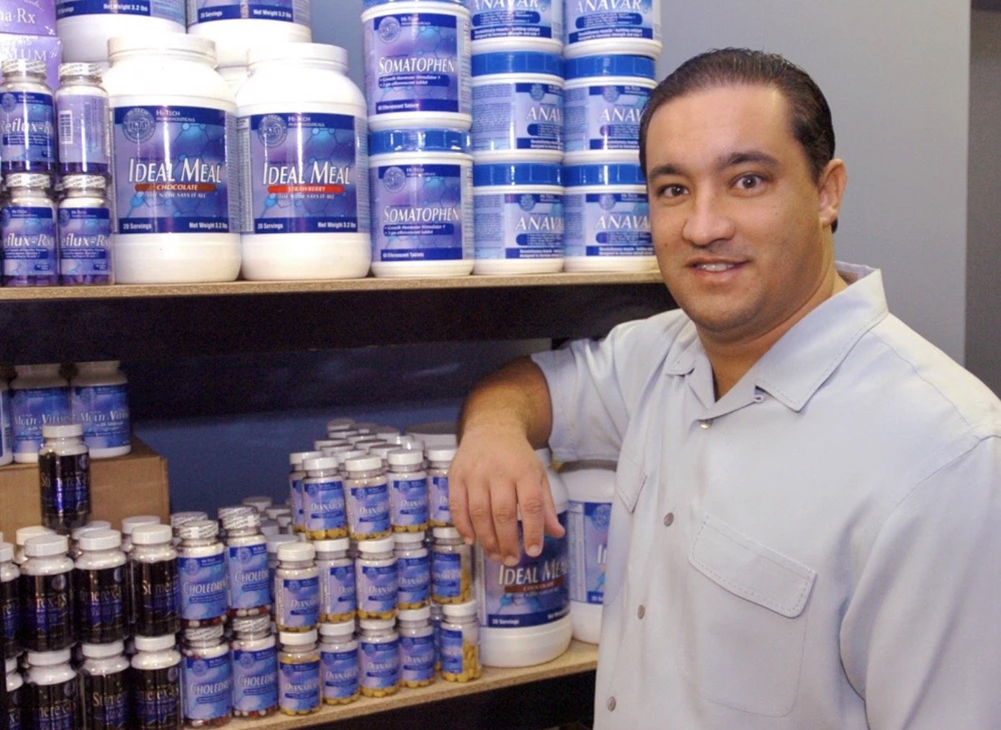 Man standing next to shelves filled with various nutritional supplements and protein powders, showcasing a health and wellness store environment.