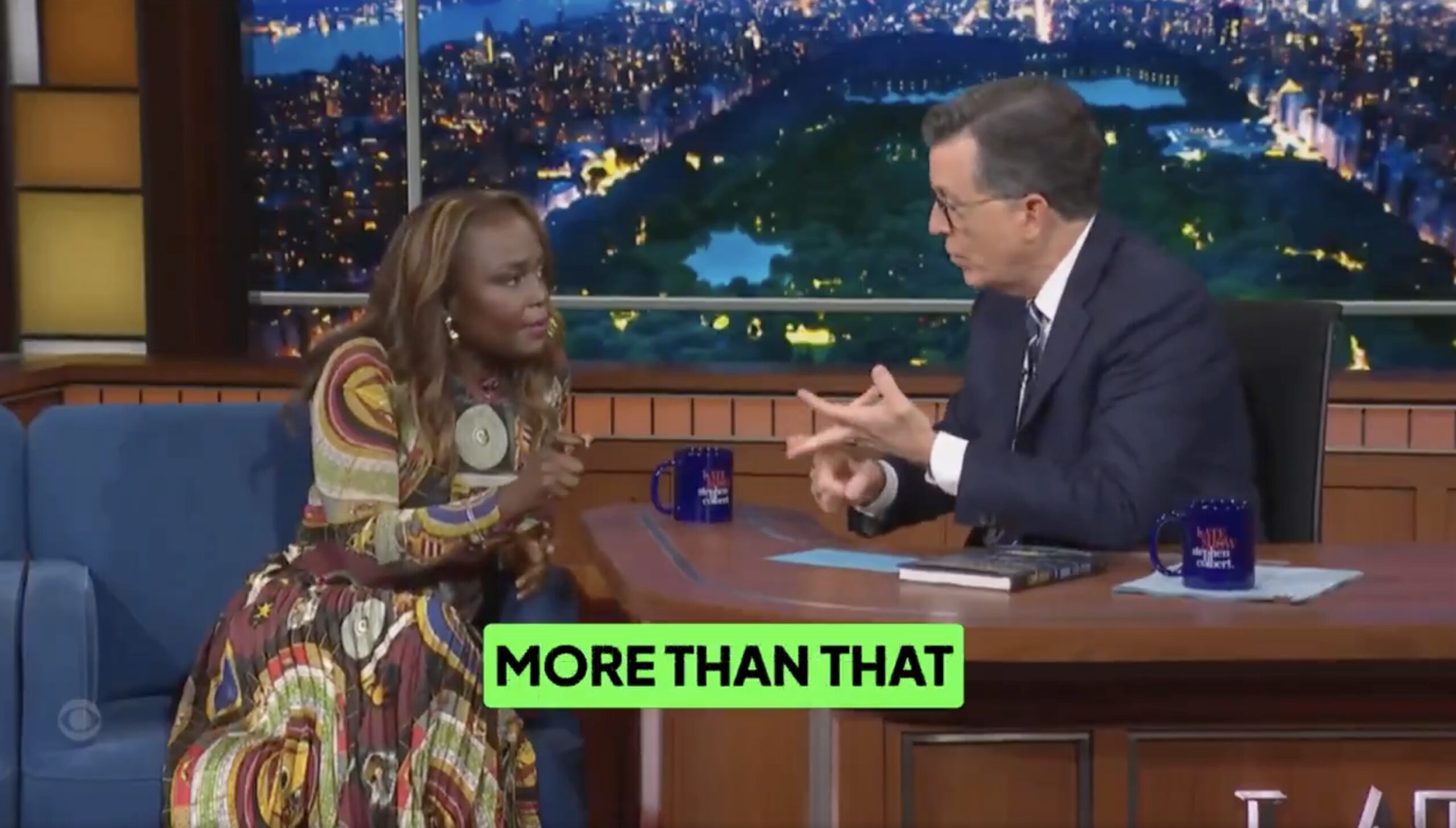 Talk show host Stephen Colbert engages in an animated discussion with a guest, set against a vibrant cityscape backdrop.