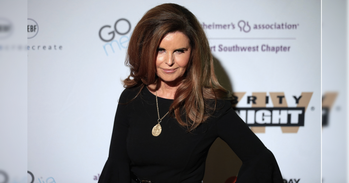A woman with long hair wearing a black dress and a gold pendant smiles confidently at a charity event backdrop.