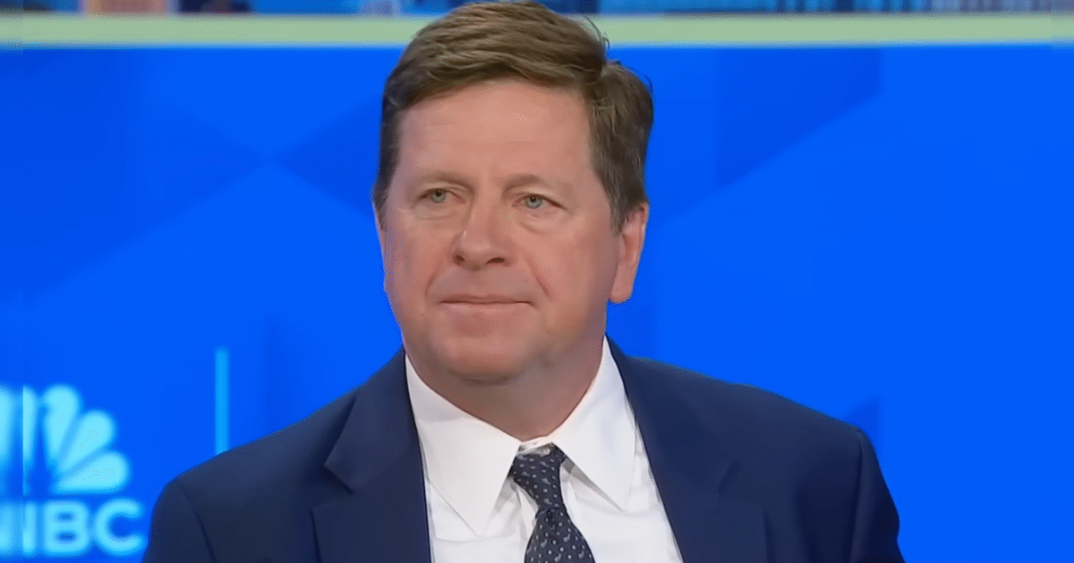 Business professional with short hair and a suit, sitting in front of a blue background during a television interview.