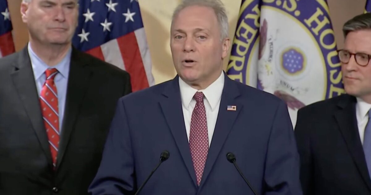 U.S. House Speaker addresses the media during a press conference, flanked by colleagues and American flags in the background.