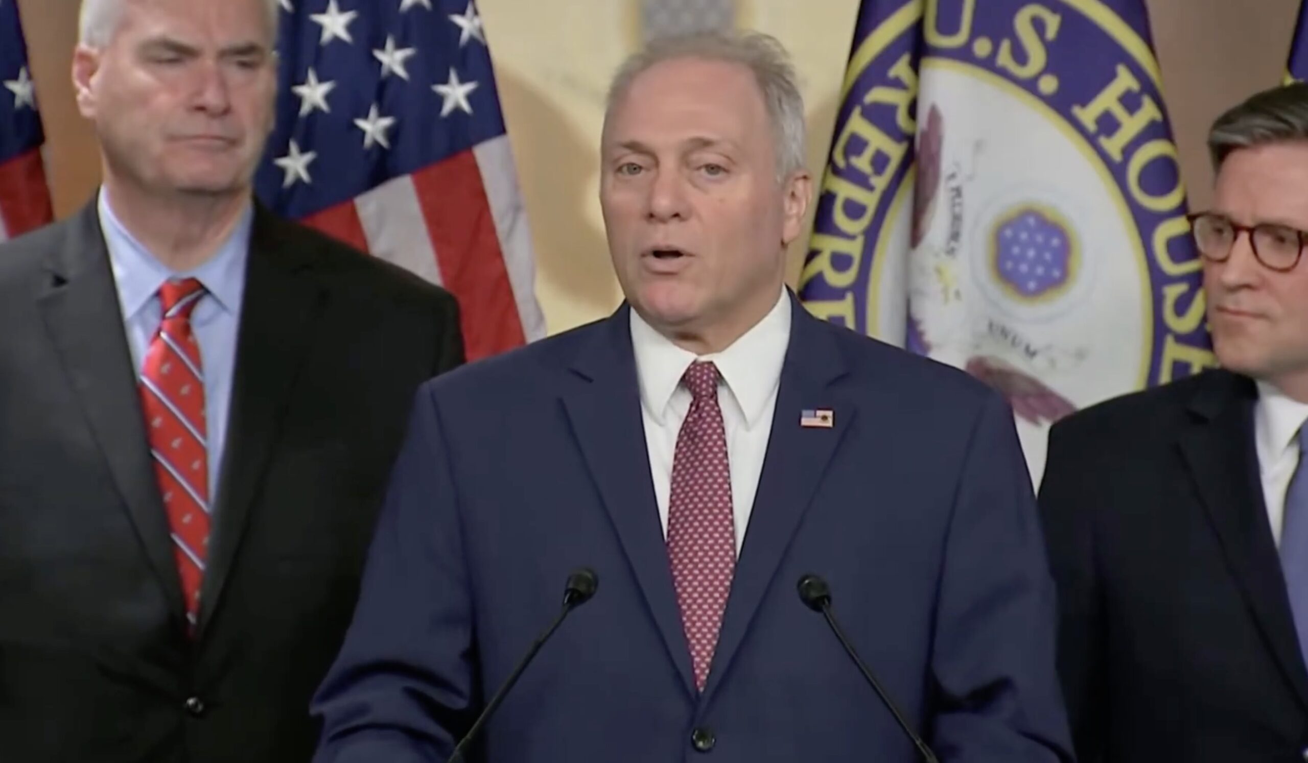U.S. House Speaker addresses the media during a press conference, flanked by colleagues and American flags in the background.