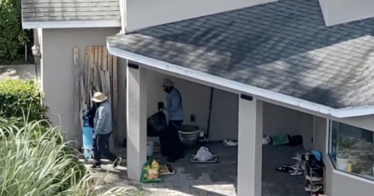 Two individuals working outside a home, one carrying a trash bag and the other with a blue container, surrounded by various items on the ground.