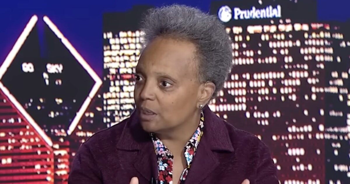 A woman with short gray hair speaks passionately in front of a city skyline backdrop, discussing important community issues during a televised interview.