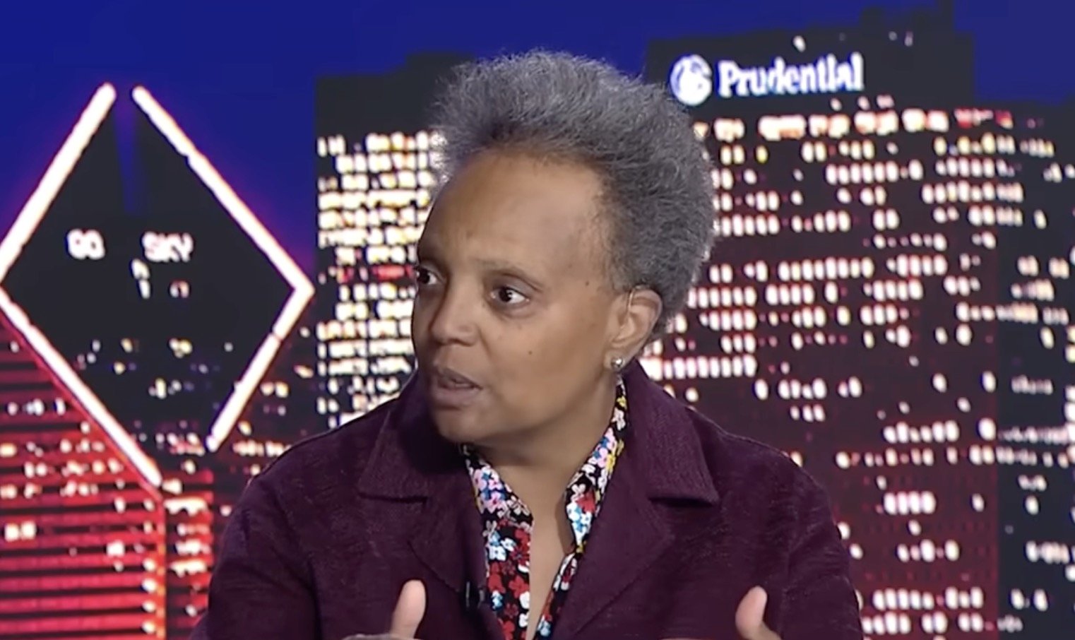 A woman with short gray hair speaks passionately in front of a city skyline backdrop, discussing important community issues during a televised interview.