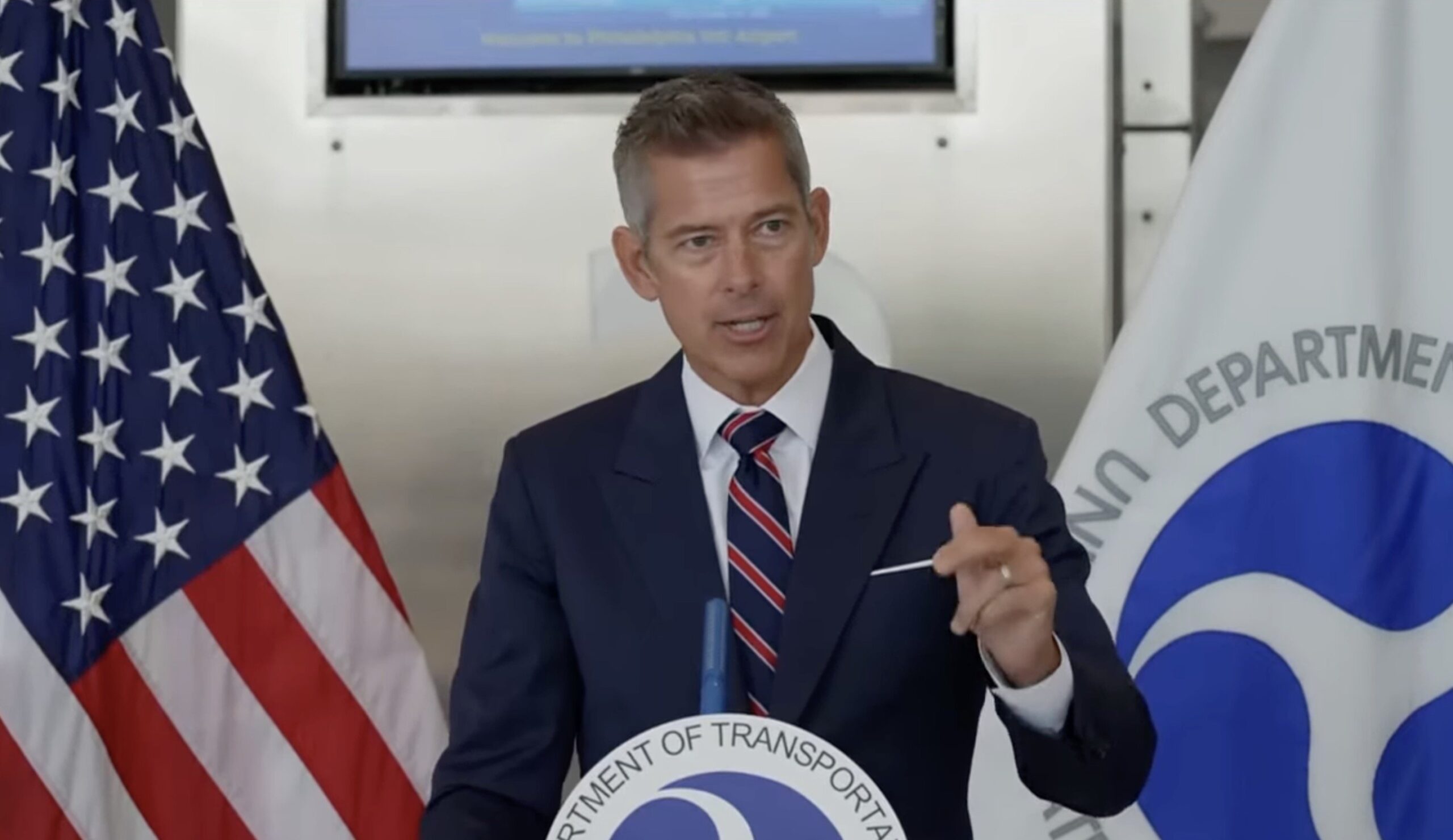 Man in a suit speaking at a podium with the Department of Transportation logo, flanked by an American flag, during a press event.