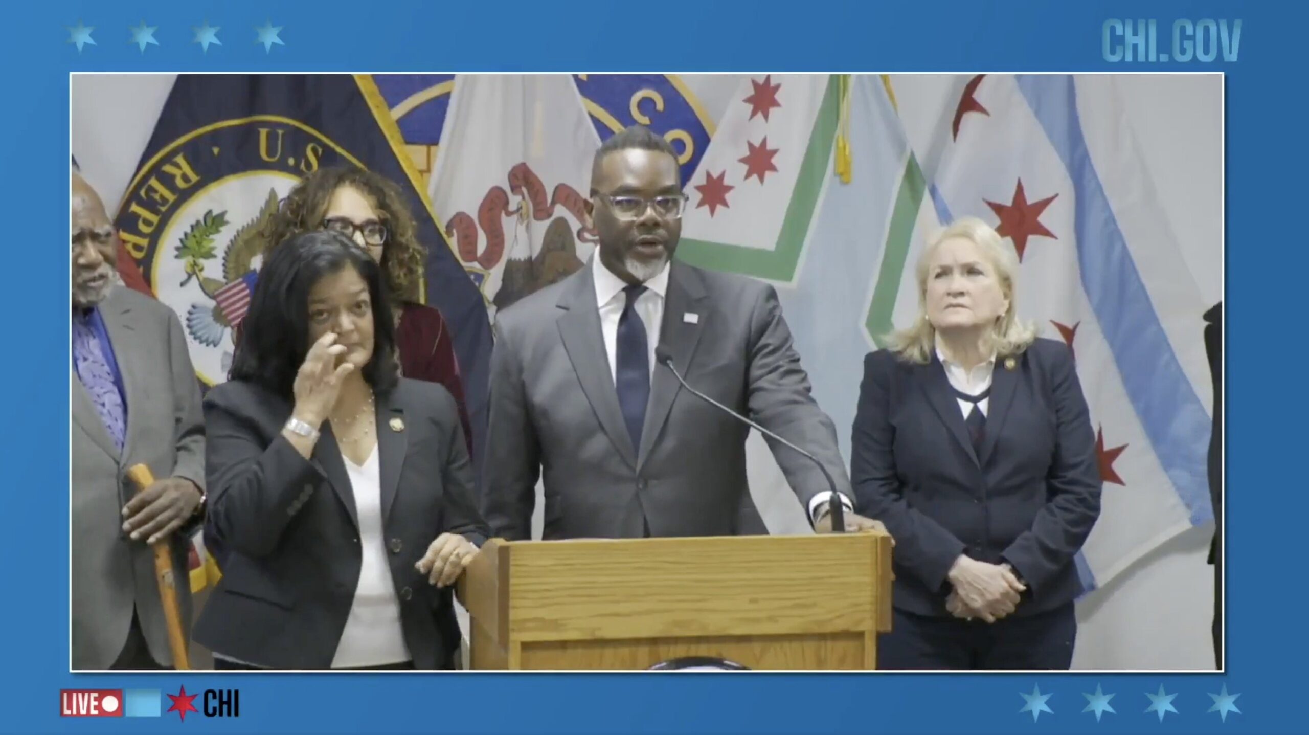 Press conference featuring Chicago officials discussing community initiatives, with multiple flags in the background and speakers at a podium.