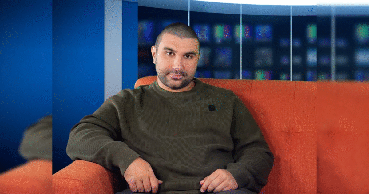 Man sitting on an orange couch in a studio setting, wearing a green sweater, with a blue background featuring a television screen.