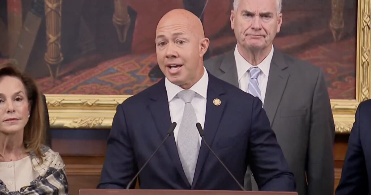 A speaker addressing an audience during a formal event, flanked by colleagues in a historic room with ornate decor.