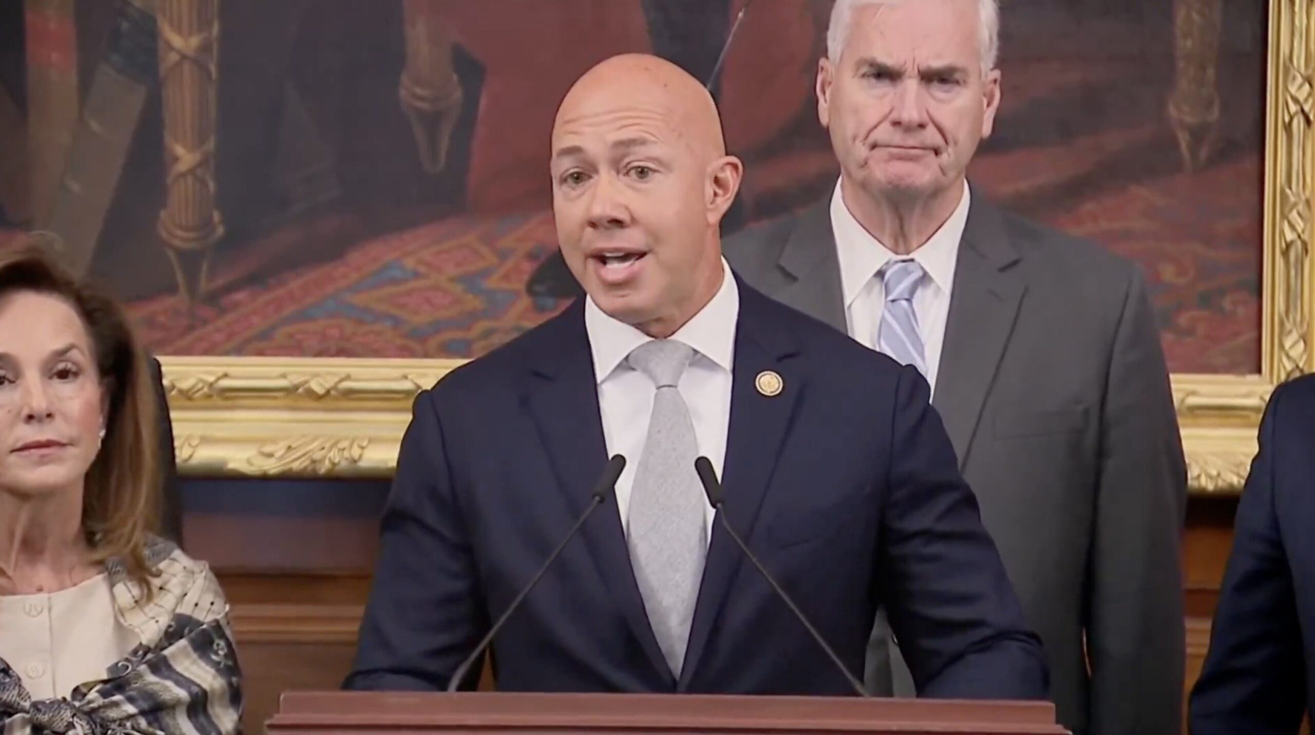 A speaker addressing an audience during a formal event, flanked by colleagues in a historic room with ornate decor.
