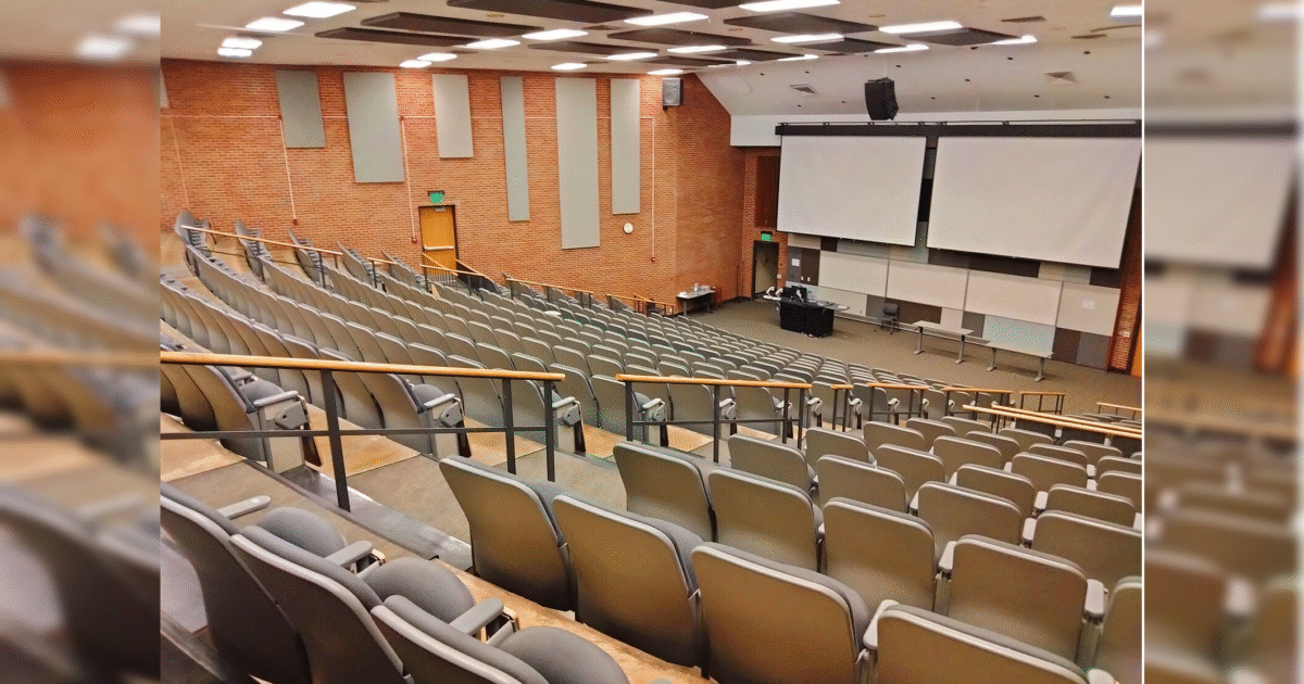 A spacious lecture hall featuring tiered seating, large projection screens, and a central teaching area, suitable for educational presentations and classes.