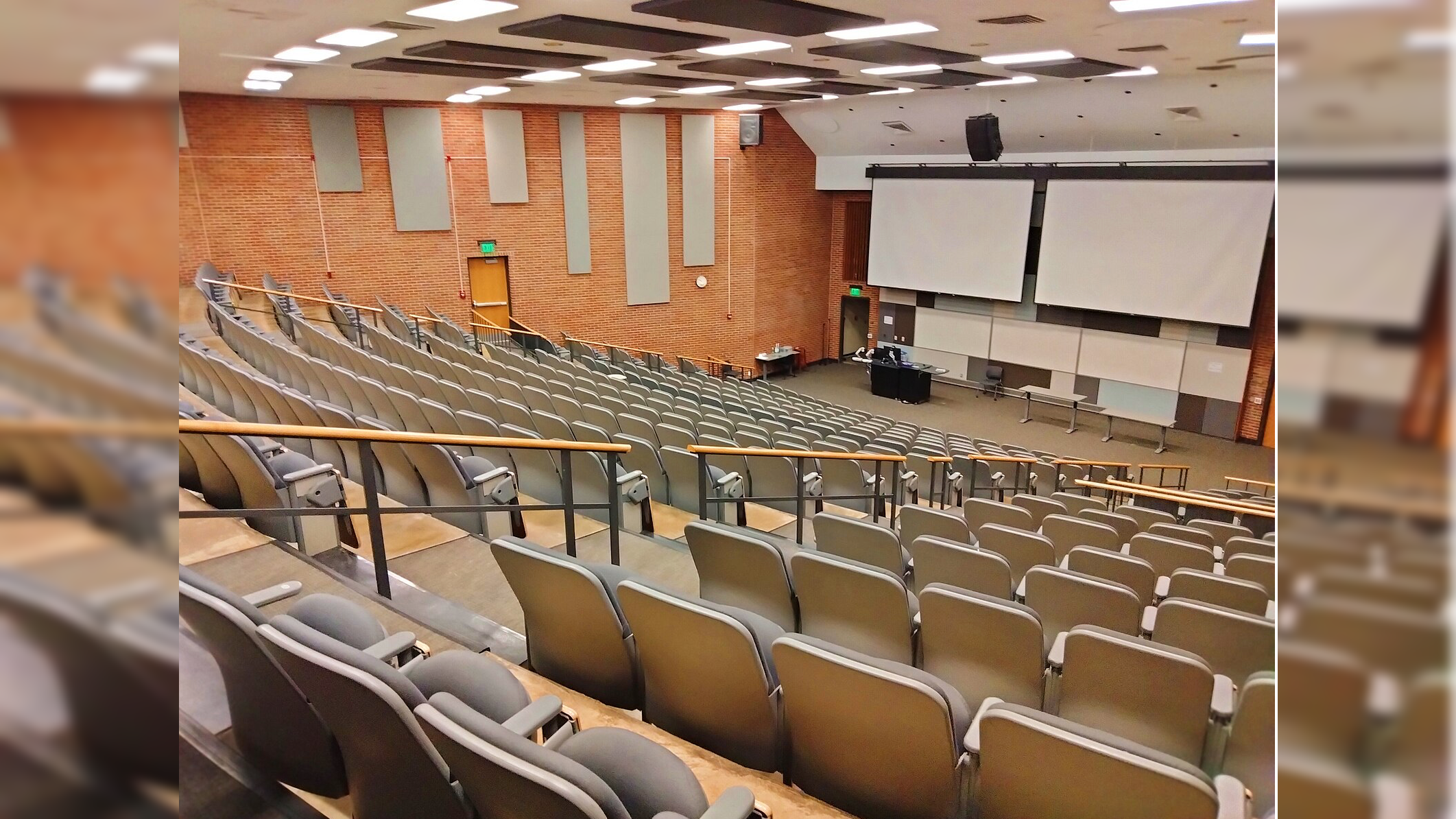A spacious lecture hall featuring tiered seating, large projection screens, and a central teaching area, suitable for educational presentations and classes.