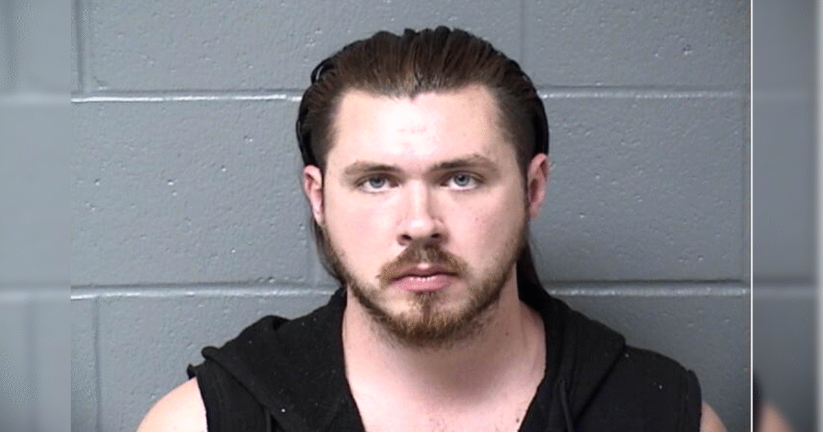 Mugshot of a male individual with long hair and a beard, wearing a sleeveless black hoodie, against a gray wall backdrop.