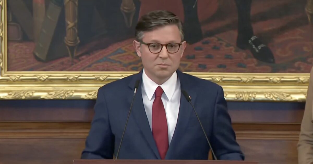 Man in a blue suit and red tie delivers a speech at a podium, with a historical painting in the background.