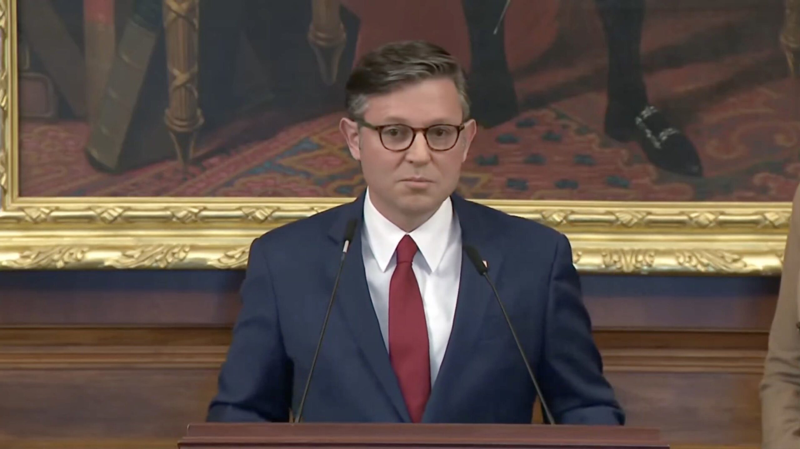 Man in a blue suit and red tie delivers a speech at a podium, with a historical painting in the background.