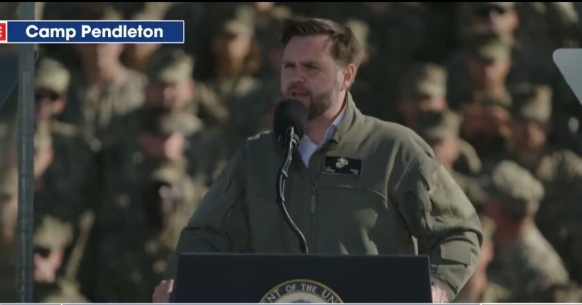 Speaker addressing military personnel at Camp Pendleton during a live event, with service members visible in the background.