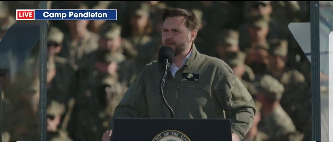 Speaker addressing military personnel at Camp Pendleton during a live event, with service members visible in the background.