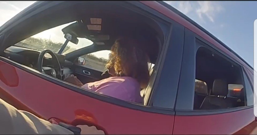 A person sitting in a red car, looking out the window, with a scenic background and bright sunlight illuminating the scene.