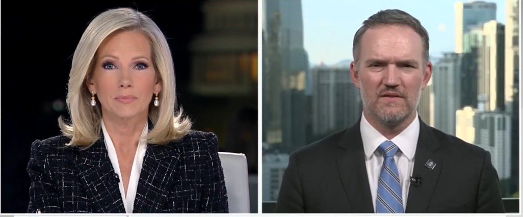 News anchors discussing current events in a studio setting, featuring a female host on the left and a male guest on the right with a city skyline backdrop.